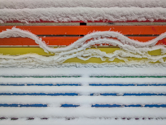 A vibrant, multi-colored wooden bench partially covered in fresh snow. The bench features horizontal slats painted in the colors of the rainbow, creating a striking contrast against the white snow.

On the upper slats, the snow has begun to slide, forming elegant, wave-like curves and "ribbons" rather than a solid blanket.

A rainbow bench is a bench in a public space that is painted in the colors of the rainbow flag of the LGBT+ movement. Its function is similar to that of a rainbow crosswalk.