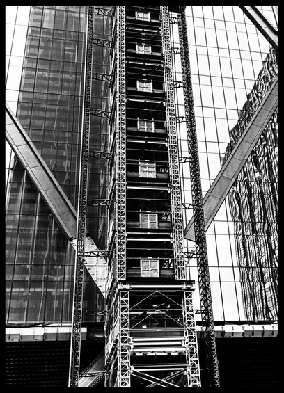 A black and white photo that shows an outside elevator going down the middle. Then crisscrossed beams coming up on the side and a dark opening for each floor with small lights going across. On the right side is a glass windowed high rise. Another building from across the street is being reflected onto the bottom of that and looks very wavy. There are two large beams going out at angles from the elevator one a bit higher than the other. One is going to the upper right hand side and the other to the upper left-hand side. Behind the left side is a dark windowed building. The bottom part of the photo shows a ledge going through and it is dark underneath on the right and left sides. It all combines to make an interesting geometric patterned photo, 🤞 