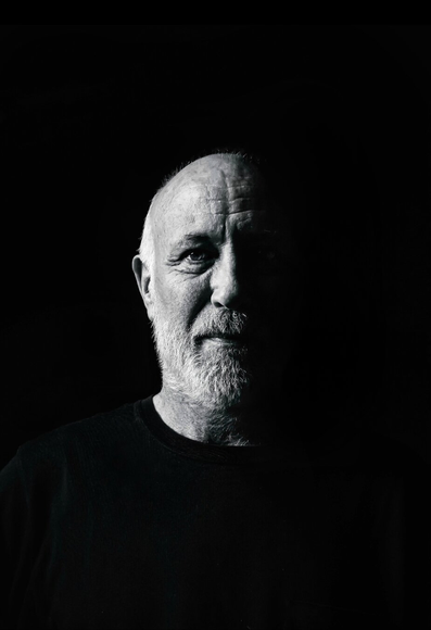 This black and white portrait features a man ( my better half) with a white beard partially illuminated against a stark black background. He faces the camera, with the left side of his face and his gaze clearly visible, while the right side is shrouded in shadow. It is a from the shoulders up view. His beard is full. He is wearing a dark, plain t-shirt.