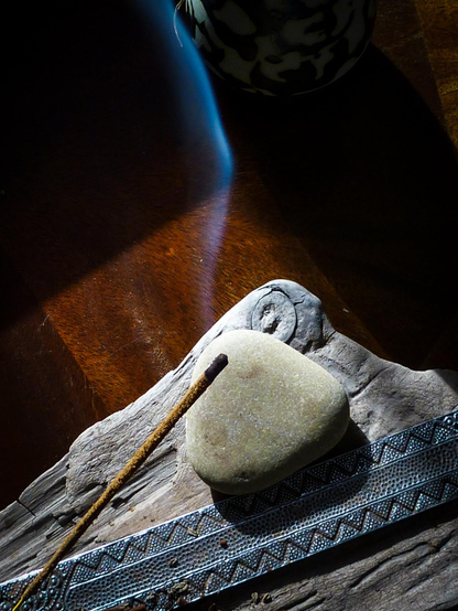 Looking down onto a stick on my incense burning in an ornate metal holder. Sitting on a dark wood table the holder sits on a flat piece of drift wood and a smooth rock sits nearby.