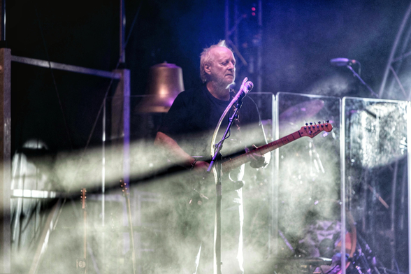 Das Bild zeigt einen Musiker der in der mitte einer bühne steht und eine E-Gitarre spielt. Der Gitarrenhals wirft einen langen Schatten im Bühnennebel.
Er trägt ein schwarzes T-Shirt und hellere Hosen. Vor ihm steht ein Mikrofonständer, und er blickt nach oben und leicht nach rechts. Die Bühne ist in dunkle Töne getaucht, mit intensivem blauem und violettem Bühnenlicht, das von oben und hinten durch den aufsteigenden Rauch oder Nebel filtert. Im Vordergrund und Hintergrund sind verschiedene Bühnenelemente wie Metallgerüste, transparente Trennwände, ein Schlagzeug und andere Instrumente teilweise sichtbar, allesamt verschwommen durch den Nebel und die Beleuchtung.