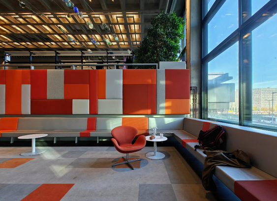 A photo showing a modern interior space with large windows overlooking a city. We look at a small seating area with small round table and swivel chair and modular orange and gray wall panels.