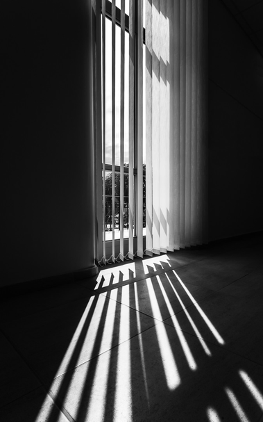 Fenster bis zum Boden, Innenansicht. Vertikalrollos, kräftiges Sonnenlicht von außen. Scharfe, perspektivische Schatten durch die Rollos auf dem Boden.