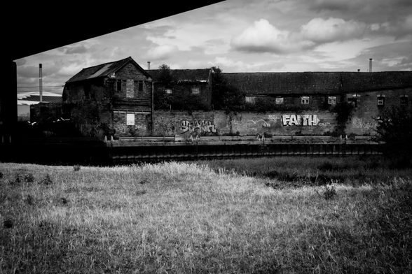 A black and white image of an old industrial building, covered in graffiti, along the quay of a river shot from underneath a road viaduct, which creates some negative space in the top left.