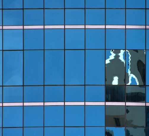 das quadratische bild zeigt eine nahaufnahme einer fensterfront eines hochhauses. die fensterelemente sind aus glas oder einem reflektierenden metall. sie sind geteilt durch dunkle linien, die ein gittermuster ergeben. zwei weiße linien, eine oben, die andere unten, verlaufen horizontal durch das bild.
fast alle fensterelemente reflektieren blauen himmel. nur rechts unten sind auf einigen der elemente reflektionen von anderen gebäuden zu sehen. diese sind durch das fenstermaterial verzerrt.