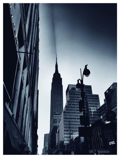 A moody monochrome photo in slightly blue tones, showing a street-level view of the Empire State Building as seen from a few blocks away on 34th Street in Manhattan. The building is backlit by the sun, and the spire casts a long dramatic shadow across the sky. In the foreground at left is the facade of the Macy’s building; at right is an antique double streetlight with gumball luminaires and some high-rise buildings across the street.