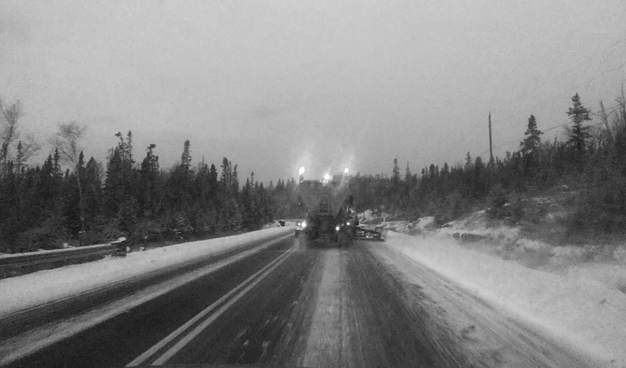 Black and white photograph taken from a car driving behind a plow on a road that is half-covered in snow and ice, bordered by forest on both sides and power lines on the right. The weather is dark, early evening, and the plow has bright headlights at the rear. The plow blade is wider than the roadway, making passing dangerous on this winding road. The image is blurred by the low light and the dirty windshield.

Photographie noir et blanc prise depuis une voiture, roulant derrière une charrue sur une route à moitié enneigée et glacée, bordée par la forêt des deux côtés et des lignes électriques à droite. Le temps est sombre, en début de soirée, et la charrue a des phares lumineux à l'arrière. La gratte est plus large que la voie, rendant les dépassements dangereux sur cette route pleine de courbes. L'image est rendue floue par la lumière faible et le pare-brise sale.