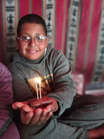 Elias, wearing glasses and a gray hoodie, smiling widely while holding a small chocolate treat with two lit candles on top; red and beige patterned curtain backdrop.
