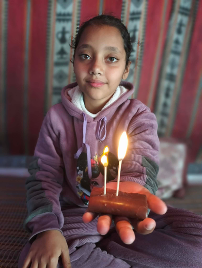 Aylan smiling, holding a piece of chocolate with two lit candles, wearing a purple hoodie, background of patterned fabric.