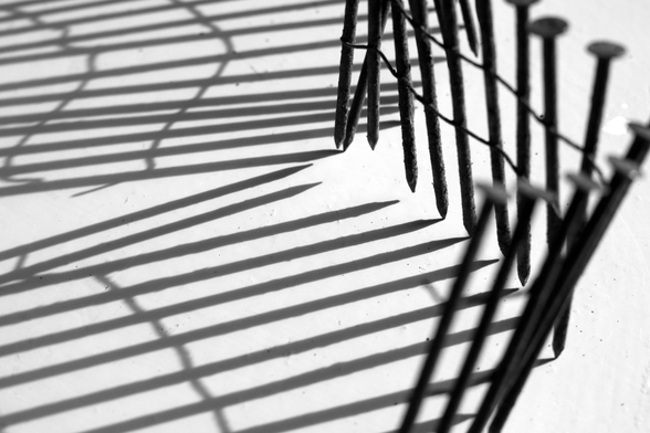 Schwarzweiße Nahaufnahme von Nägeln, die durch einen Draht zusammengehalten werden und wie ein Holzzaun aufrecht stehen. Das Licht kommt von rechts, die Nägel werfen lange Schatten.

Black and white close-up of nails held together by a wire and standing upright like a wooden fence. The light comes from the right, casting long shadows from the nails.