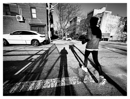 Black and white photo of a city street intersection, lit by sunlight from behind the camera. At center frame is my long shadow stretching towards the sidewalk at the far end. To the right is a jogger in mid-stride facing away from camera, their own long shadow alongside mine. Their head and upper back are darkened by the shadow of some structure out of frame. In the background are parked cars, low apartment buildings, trees, and a clear sky.