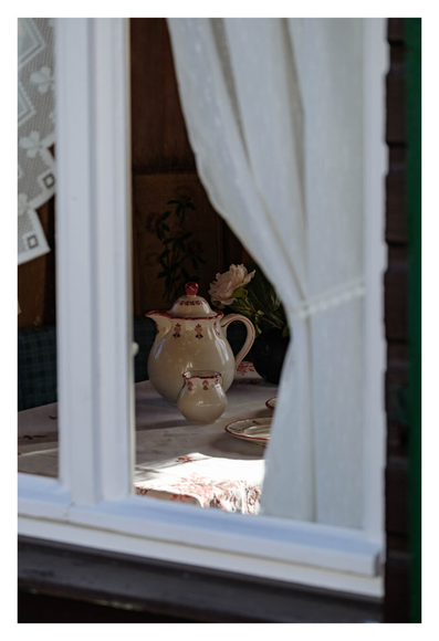 Foto im Hochformat. Teleaufnahme, Blick durch ein Fenster mit weißem Fensterrahmen. Ein weißer Vorhang ist rechts festgebunden und bildet so eine leichte Kurve. Zeichen diesem Vorhang und dem mittleren Balken im Rahmen wid der Blick freigegeben auf einen gedeckten Tisch. Eine cremefarbene Teekanne mit roten Rändern, ein kleines Milchkännchen und ein Teller im gleichen Design. Teile einer Blumenvase mit Blumen sind auch zu sehen. Die helle Tischdecke mit gestickten Blumen darauf passt zum Gesamtbild.der Tisch und Teile des Geschirrs werden von der Sonne durch ein anderes Fenster beschienen. Es ist das Fenster einer unbewohnten kleinen Gartenlaube am Wegesrand.