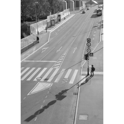Black and white street photography taken in Luxembourg city showing a big avenue from above, with someone waiting to cross it. The winter sun casts big shadows on the street, from the top right corner.