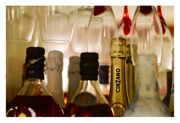 Several bottles are clustered together, including dark liquor bottles in the foreground and a prominent gold-foil-wrapped bottle labeled "CINZANO" in the center. Above the bottles, empty wine glasses hang upside down from a rack.