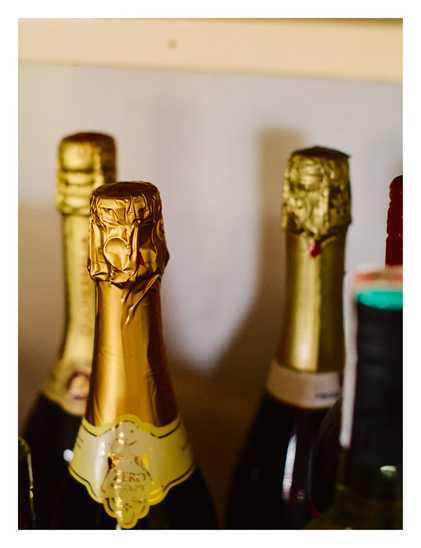 The foil-wrapped necks of several bottles, likely champagne or sparkling wine. The image features a shallow depth of field, highlighting a bottle in the left foreground with crinkled gold foil and a yellow neck label. Behind it, two other gold-foiled bottles and a dark bottle neck on the far right are visible but soft and out of focus, set against a plain, light-colored background.