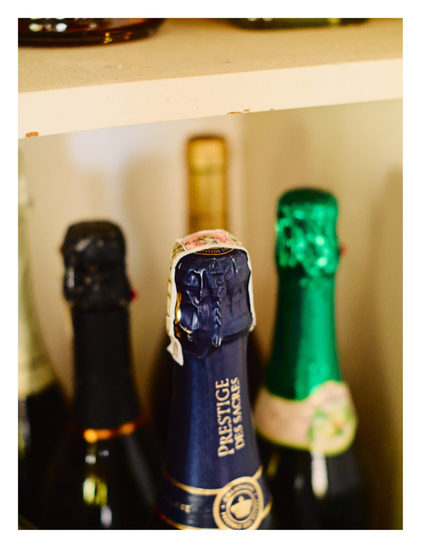 Several champagne or sparkling wine bottles stored on a shelf. The image focuses sharply on the central bottle, which features a dark blue foil neck and a label reading "PRESTIGE DES SACRES." Flanking the center bottle are two out-of-focus bottles—one with black foil on the left and one with bright green foil on the right—while a blurry gold bottle neck is visible in the background beneath a shelf.