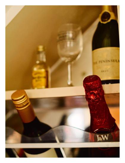 A bottle in the foreground wrapped in bright red crinkled foil with a label reading "J&W." To its left, an angled bottle with a gold screw-top cap is visible behind a clear plastic guard. The background is out of focus, showing a top shelf containing a bottle of sparkling wine labeled "THE PENINSULA," an empty wine glass, and a small bottle of amber liquid.
