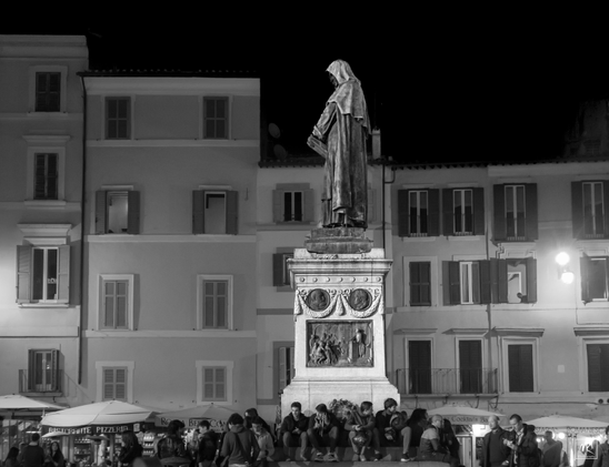 Nighttime black and white photo of the statue of Giordano Bruno in the plaza Campo d’Fiore, Rome, which the site of his execution for heresy - burned at the stake - by the Roman Inquisition. The statue is seen from its left side and there is a crowd of young adults sitting around its base.