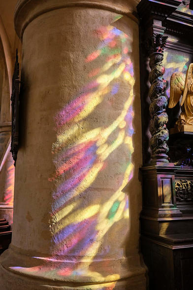 multicolored patterns from a large stained-glass window are projected by the sun onto a large stone column (and intricately carved woodwork) in a medieval cathedral. 