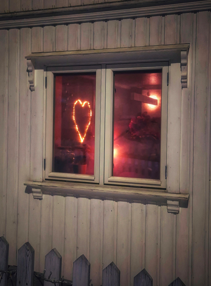 A window of an older wooden house. Inside a heart shaped light hangs on the wall. The light in the rest of the room is rather purple-red.