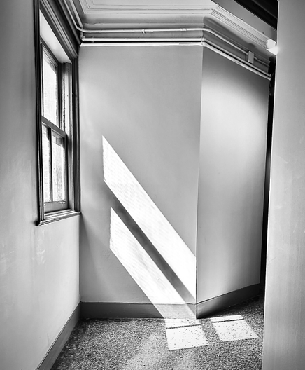 Black and white photo taken in the hallway of a pre-war office building. The floor is old dirty stone of some sort. The walls are gray with darker gray trim. There is a square window on the left-hand side. Shooting down from the window in the shape of long parallelograms are two sun shadows hitting the wall and floor