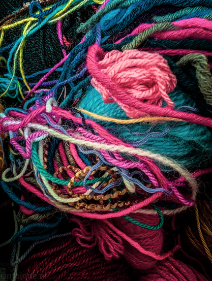 A vibrant, close-up of a tangled collection of multi-colored yarn. The composition is dense and chaotic, filled with various textures, weights, and fibers overlapping in every direction.

The image is dominated by a rich, saturated color scheme. Deep teals, bright magentas, and hot pinks. Also lime green, soft lavender, and earthy forest greens. A strand of gold-toned, beaded trim adds a distinct glint and different texture to the center-left.