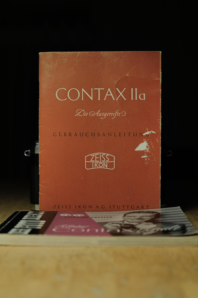 Digital color photo of a worn red instruction booklet reading “CONTAX IIa,” “Die Ausgereifte,” and “Gebrauchsanleitung,” with a Zeiss Ikon logo and “Zeiss Ikon A.G. Stuttgart” at the bottom, standing upright on a wooden table with a Contax camera partially visible behind it against a dark background. (Photo by me, CC-BY-4.0)