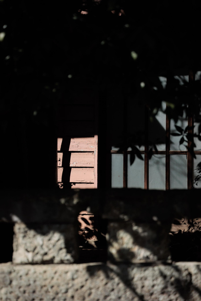 A glimpse of a rustic house partially obscured by green foliage and a stone wall in bright sunlight.