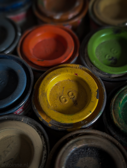 A close-up shot of several small, weathered paint tins used for model building. The composition is tightly packed, focusing on the textured lids of the cans. 

A vibrant yellow lid sits in the center, featuring the number "69" embossed in the metal. The paint on this lid is chipped and worn, showing a dark, grimy patina.

 Surrounding the central yellow are lids in bright orange, lime green, deep navy blue, and a muted tan. The colors are saturated but darkened by shadows and age.