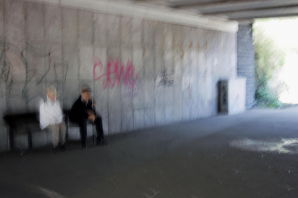 Eine Frau mit weißer Jacke und ein Mann ganz in schwarz gekleidet sitzen auf einer Bank in einer Unterführung. Die Wand ist zum Teil mit rotem Graffiti bemalt. Am Ausgang erkennt man grünliche Büsche. Das Foto ist mit Absicht verwackelt und verwischt.

A woman wearing a white jacket and a man dressed entirely in black are sitting on a bench in an underpass. The wall is partially covered in red graffiti. Greenish bushes can be seen at the exit. The photo is deliberately blurred and out of focus.