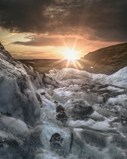 Golden sunlight bursts over a rugged glacial landscape, illuminating textured ice formations and flowing meltwater while dark clouds frame the horizon and distant valleys stretch into the background.