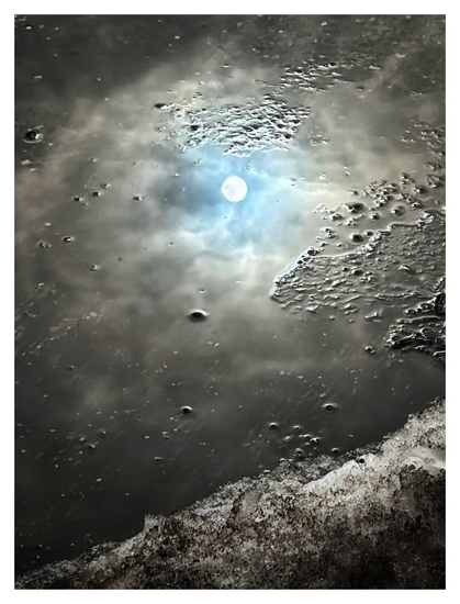 Closeup of a very shallow gray puddle on a city street. Little concrete bumps stick out of the water’s surface like islands, and a patch of dirty snow and ice slopes upward from left to right in the foreground at bottom frame. Reflected in the water is a pale winter sun, in a small opening of blue sky surrounded by gray clouds. Although it’s clear what the image is, it also makes me think of a snow-capped mountain lit by a waning sun, shining through the clouds of a gathering storm.