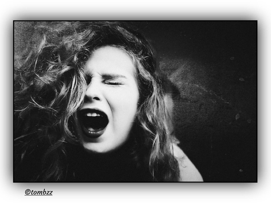 A raw, high‑contrast frame that hits with emotion. A woman’s face fills the shot, eyes shut tight, mouth wide open in a scream that refuses to settle into one meaning. It could be pain, it could be anger, it could be something tangled between the two. Light carves out the tension in her cheeks and the pull of her brow, giving every line a sense of strain.  

Her hair is long, thick, curly, pushed outward in a wild, uncontained motion, as if caught in the middle of a sudden burst of feeling. Each curl catches the light differently, creating sharp, shifting highlights.  

The background is dark and rough, a textured surface that adds to the unease. The whole image feels charged, unresolved. It doesn’t explain itself. It leaves you standing in the space between sound and silence, with questions that don’t settle.