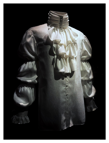 Prince’s white silk shirt from his 1980s Purple Rain era, with puffed sleeves and a high collar with a fall of ruffles, on display in a museum exhibit against a black backdrop. Photo taken at the recent exhibition “Superfine: Tailoring Black Style” at the Metropolitan Museum of Art in NYC.