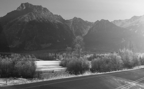 A black and white photograph of a winter mountain landscape, taken from a car on the road. High mountains with sharp peaks are visible in the background, their slopes covered in snow. At the foot of the mountains, in the middle of the photo, are trees covered in frost and snow-covered fields enclosed by a fence. The road is visible at the bottom of the image. The dirty windshield and the bright sunlight make the photo slightly blurry.

Photographie noir et blanc d'un paysage montagneux en hiver, pris à bord d'une voiture sur la route. De hautes montagnes aux pics acérés au soleil sont à l'arrière, avec de la neige sur leurs flancs. Au pied des montagnes au milieu de la photo se trouvent des arbres couverts de frimas et des champs enneigés et entourés d'une clôture. La route est visible dans le bas de la photo. Le pare-brise sale et la lumière éblouissante du soleil rendent la photo un peu floue.