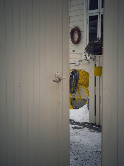 Looking through a slightly open gate into a backyard café that is only open in summer. A yellow bicycle and a yellow flower pot are the only colors. The gate and the wall of the house are all beige. A black extension cord hangs from the handlebars of the bicycle, making the picture even more dreary.
