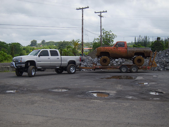 A silver pickup truck is towing a flatbed trailer with mud-covered, older pickup truck with large off-road tires.
