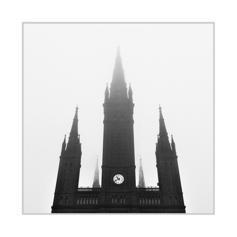Square black-and-white photograph. A large church façade is seen from a low angle as a dark silhouette against a bright, foggy sky. A tall central spire rises from the middle, flanked by two smaller towers with pointed spires on the left and right, while additional faint spires appear in the mist behind. Near the lower center of the building, a round clock face is clearly visible, providing the brightest detail within the dark architecture.