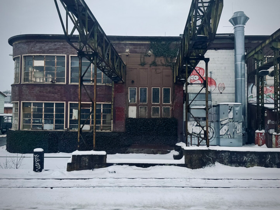 Ein verlassenes Industriegebäude mit großen Fenstern und einer geschwungenen Fassade, teilweise mit Efeu und Schnee bedeckt. Eine Metallstruktur erstreckt sich über den Eingang, und es gibt Graffiti-Kunstwerke an der Seitenwand. Die Szene spielt in einer winterlichen Landschaft mit Schneebedeckung