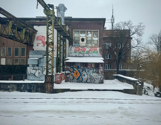 Verlassenes Industriegebäude mit Graffiti bedeckt, umgeben von Schnee. Eine Metallstruktur und Bäume sind sichtbar, mit einem bewölkten Himmel über dem Kopf.
