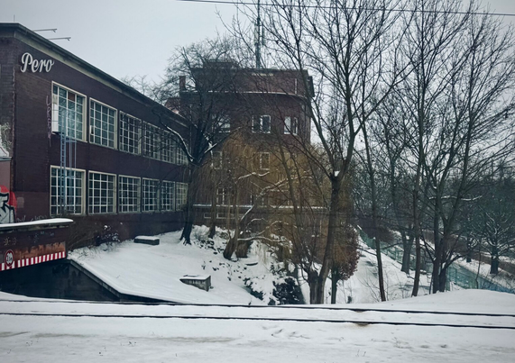 Ein Industriegebäude mit großen Fenstern, umgeben von schneebedecktem Boden und kahlen Bäumen. Die Struktur zeigt ein Schild mit dem Wort "Pero" und eine kleine Holzbank, die im Vordergrund sichtbar ist. Die Szene hat eine düstere Winteratmosphäre.