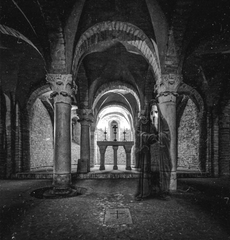 The black and white photo is of a stone crypt (San Zama) featuring a series of arched openings supported by ornate columns. At the far end, three crosses stand on top of an altar, illuminated by a light. The floors are stone-paved and worn, with a cross etched into one of the stones near the foreground. A translucent figure, wearing a long, dark robe and the white mask like that of a plague doctor, stands to the right, (recreating the face of the Spectre of Newby) almost blending into the stonework. The grainy texture and high contrast help make it feel like an old photo.