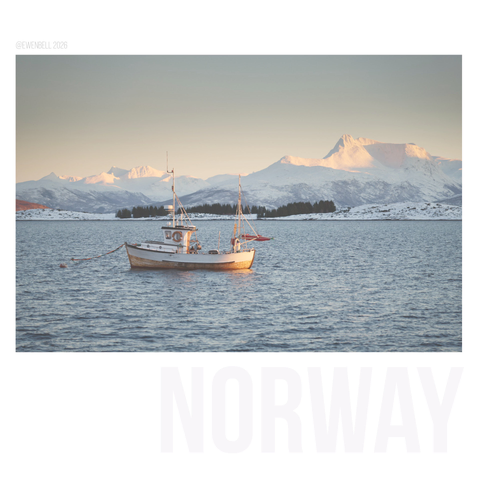 A very small fishing boat is moored in a fjord with distant mountains in the background catching a little pink light at the end of the day