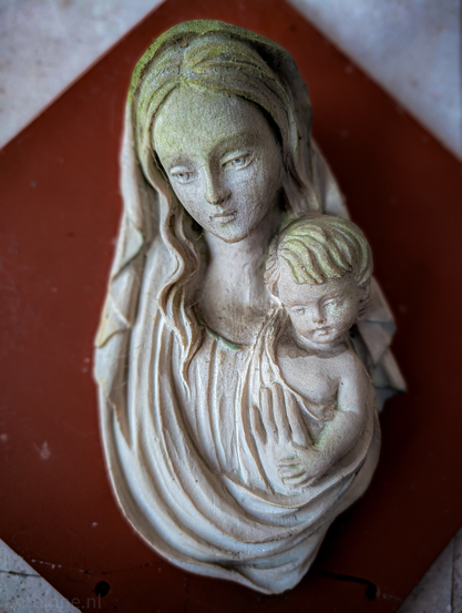 A high-relief stone carving of the Madonna and Child, capturing a serene and intimate moment. The sculpture is set against a dark, reddish-brown terracotta background, which makes the pale, weathered stone of the figures stand out. 

The Madonna is depicted with a gentle, downcast gaze and a slight, peaceful expression. Her head is tilted toward the child, and she wears a flowing veil that drapes over her hair and shoulders. The infant Jesus is nestled in the crook of her arm, his face turned slightly toward the viewer. His features are soft, and his hair is rendered in delicate, wavy tufts.
