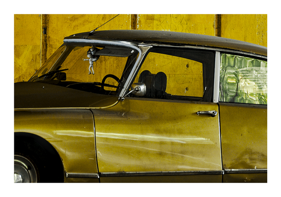 Daylight photography.
Vielle top model.
Close-up of an old yellowish car with some green reflection on a side window, parked along a yellow wall.