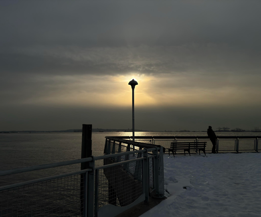 A view from a snow-covered pier, around golden hour, overlooking the waters of New York Harbor. Zigzagging metal guardrails in the foreground recede towards a tall lamppost at the center of the image. The setting sun is hidden behind the lamppost head but bathes it in a halo of light, like a miniature solar eclipse. Flanking the lamppost are the silhouettes of a wooden pillar rising out of the water and a person leaning against the guardrails. On the far horizon is the Jersey shoreline and a row of shipping cranes. A wide golden band of illuminated clouds with crepuscular rays stretches across the sky between gray clouds above and below. Taken at Valentino Pier in Red Hook, Brooklyn.