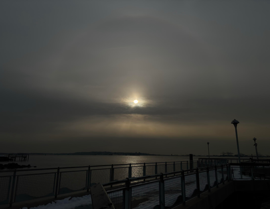 A view from a pier overlooking the waters of a wide bay. A halo of pale light arcs high over the setting sun as it shines over a strip of dark clouds. Below, golden crepuscular rays fan out in a luminous band before being obscured by more gray clouds at the horizon. Directly beneath the sun, a column of sunlight sparkles on the water. In the foreground are guardrails flanking the pier’s snow-covered walkway as it recedes to some lampposts at right frame.