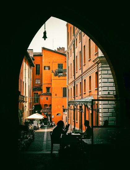 The photo is of European street scene. (Bologna) It is framed through a dark, arched stone entryway that creates a "frame-within-a-frame" effect. The photo has a shadowed foreground with two people sitting at a table talking and moves past them into a sun-drenched alleyway. The buildings in the background are warm terracotta and ochre/yellow coloured.