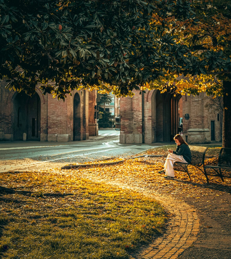 The photo is of a woman with long brown hair seated on a dark metal bench, she's looking down at her phone. She has her chin resting on her hand. She is wearing a dark green cardigan, light cream-coloured wide-leg pants, and green shoes with white stripes. The bench is positioned on a brick-paved section of the park. There are fallen leaves scattered around. A large tree with dense green and yellow foliage frames the  overhead. The ground is covered with grass and fallen leaves. In the background, there are multiple rows of old brick arches.