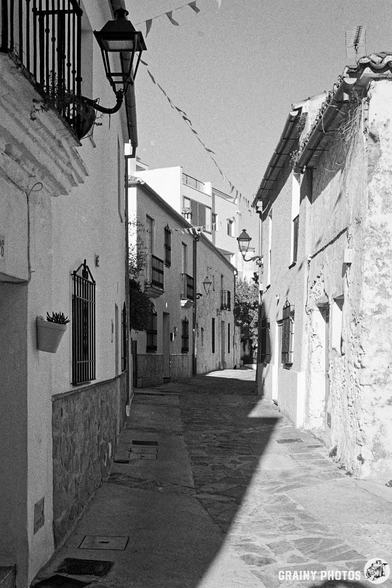 A narrow alleyway lined with whitewashed buildings, featuring a mix of balconies and stone walls. The street is illuminated by lanterns and adorned with decorative banners, creating a charming atmosphere in a quaint neighborhood.